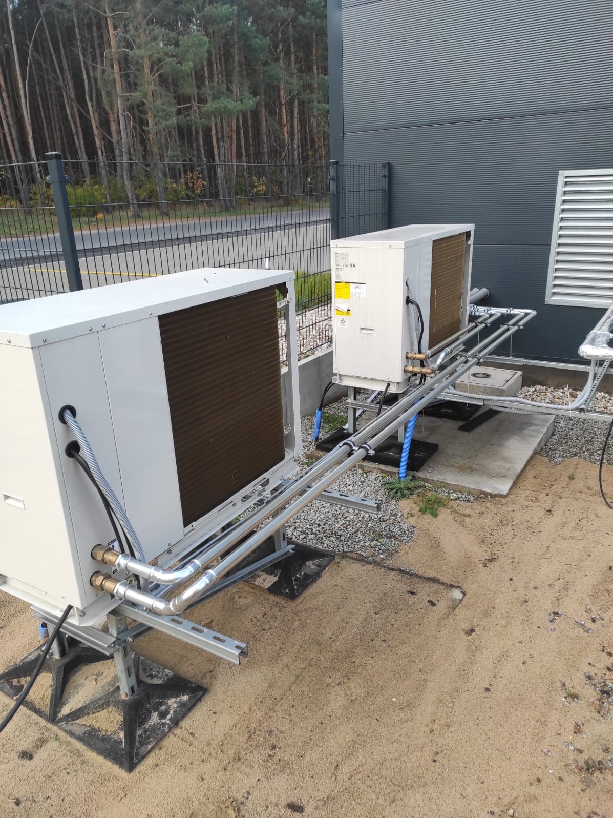 Dwie białe pompy ciepła zamontowane na zewnątrz budynku z szarą elewacją. Widoczne rury i elementy instalacji, tło stanowią drzewa i ogrodzenie.