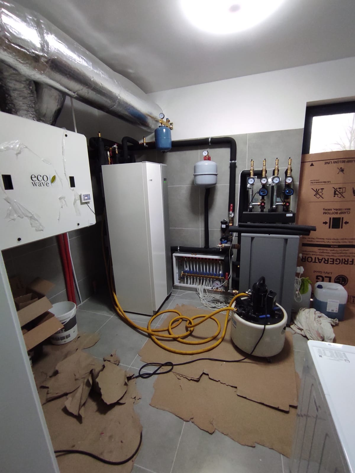 Zmontowana instalacja grzewcza z pompą ciepła Eco Wave, zbiornikami i rozdzielaczem w pomieszczeniu z szarą glazurą i kartonami na podłodze. Widoczne rury i przewody.