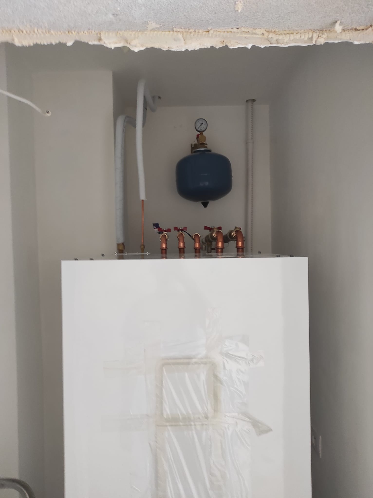 Biały piec z niebieskim zbiornikiem wyrównawczym i miedzianymi rurami w trakcie montażu w nowym budynku. Górna część pieca zabezpieczona folią.