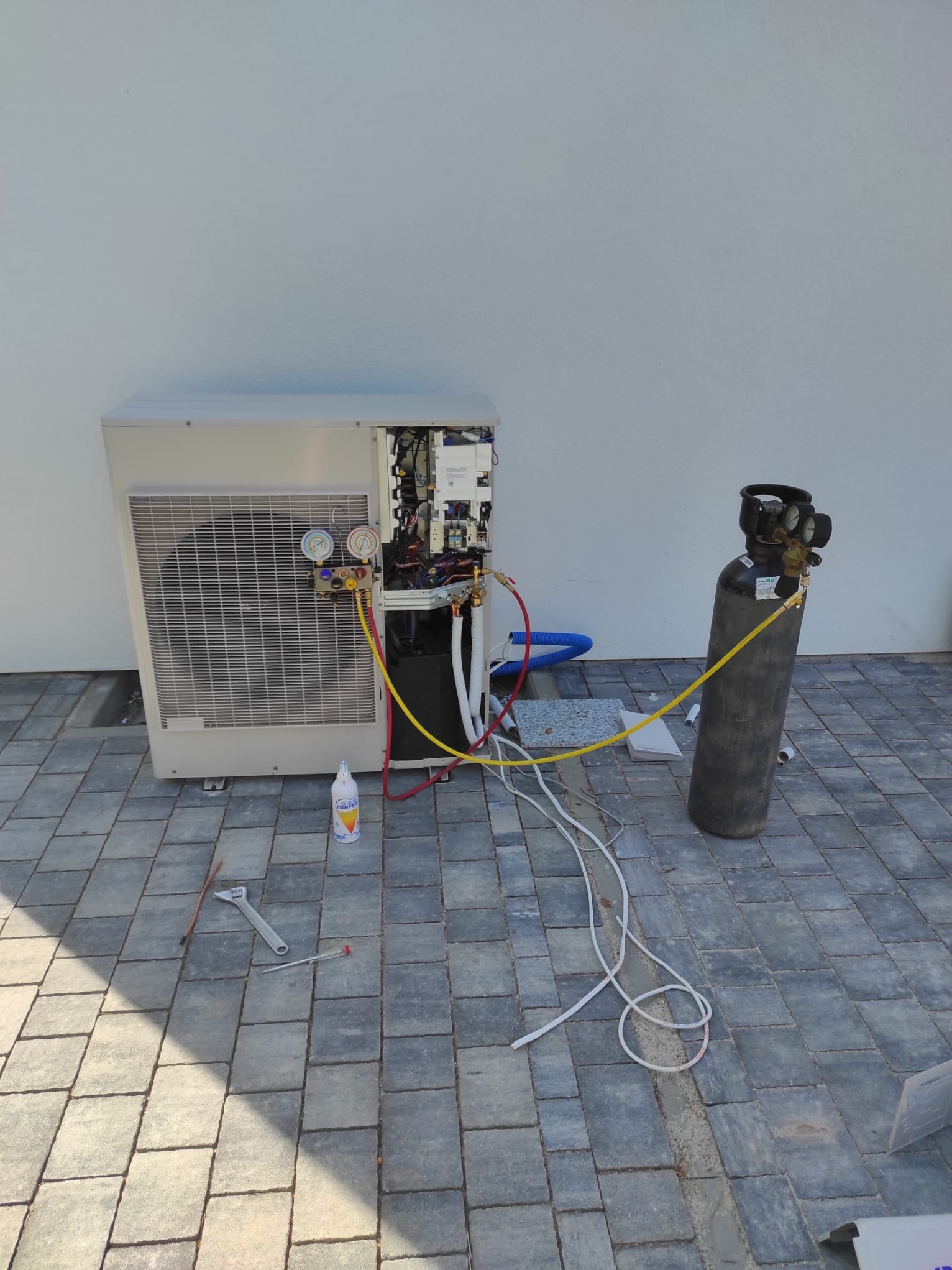 Serwis pompy ciepła: otwarty panel, manometry, butla z gazem chłodniczym, narzędzia na kostce brukowej. Prace serwisowe w toku, widoczne przewody i elementy instalacji.