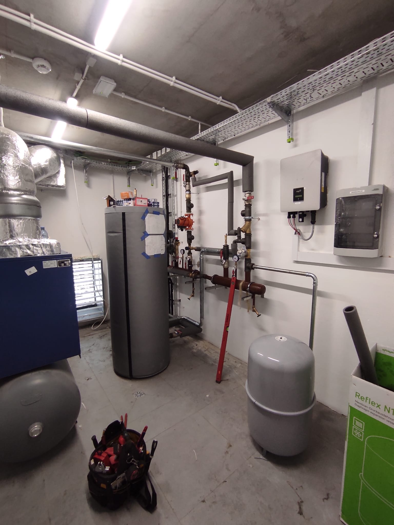 Złożona instalacja CO w kotłowni: rury, zbiorniki, armatura, pompy obiegowe i moduł solarny. Na podłodze torba z narzędziami instalatora. Widoczne elementy marki Reflex.