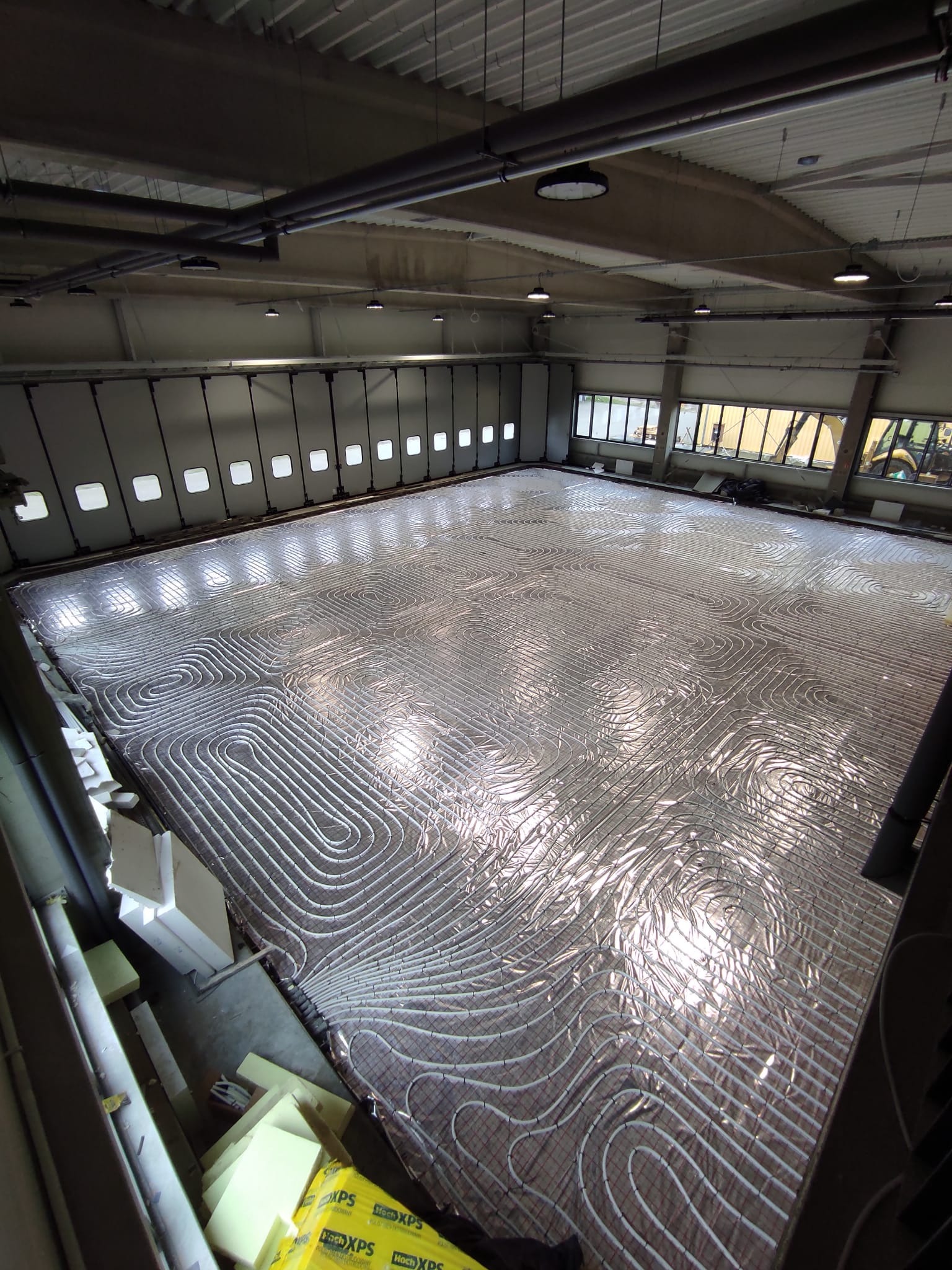 Rozległa instalacja ogrzewania podłogowego z białych rur ułożonych na srebrnej folii izolacyjnej w dużej hali przemysłowej, widok z góry.