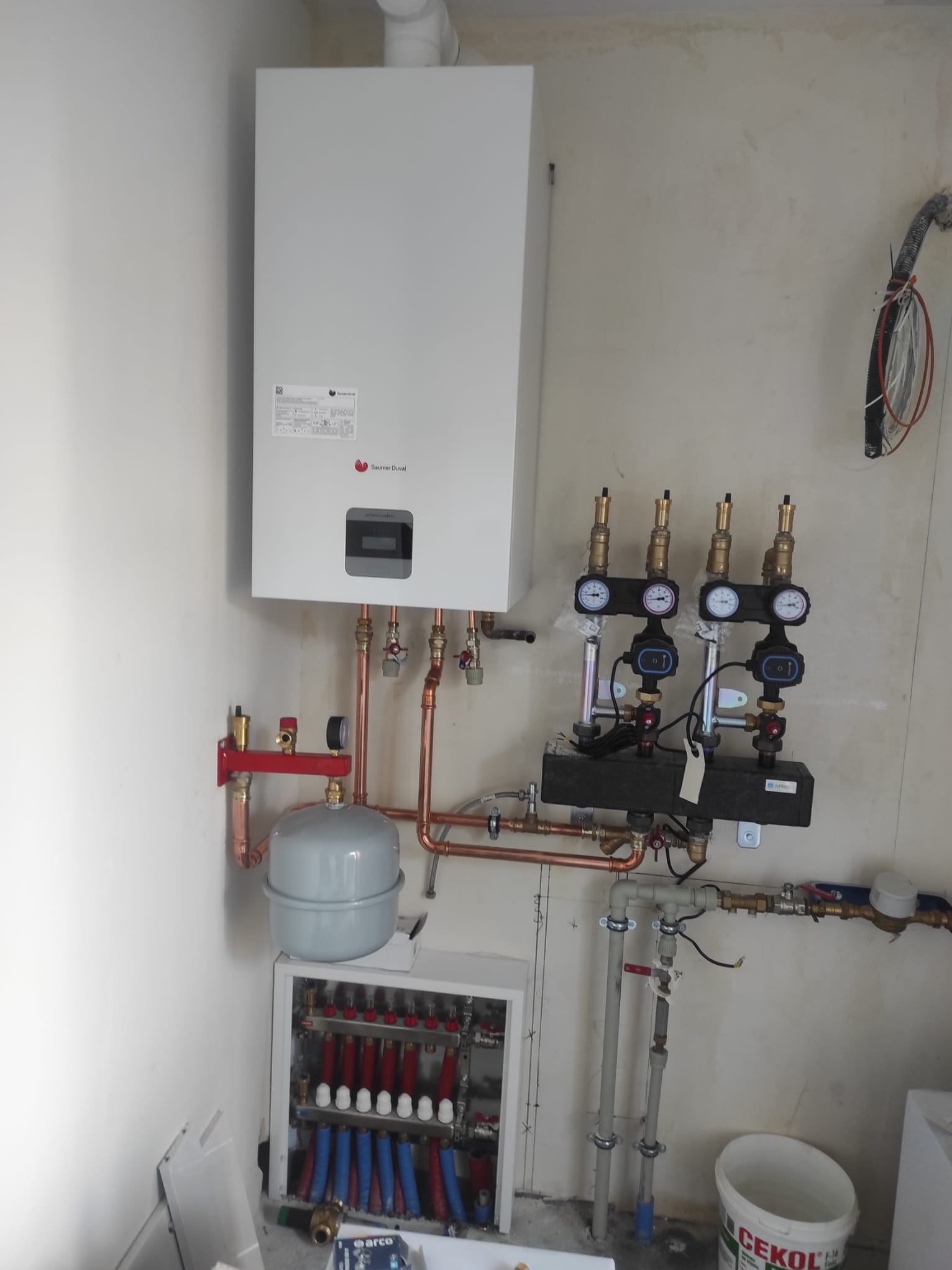 Biały piec gazowy Saunier Duval zamontowany na ścianie, z widoczną instalacją hydrauliczną z miedzianych rur i rozdzielaczem podłogowym w pomieszczeniu w trakcie wykończenia.