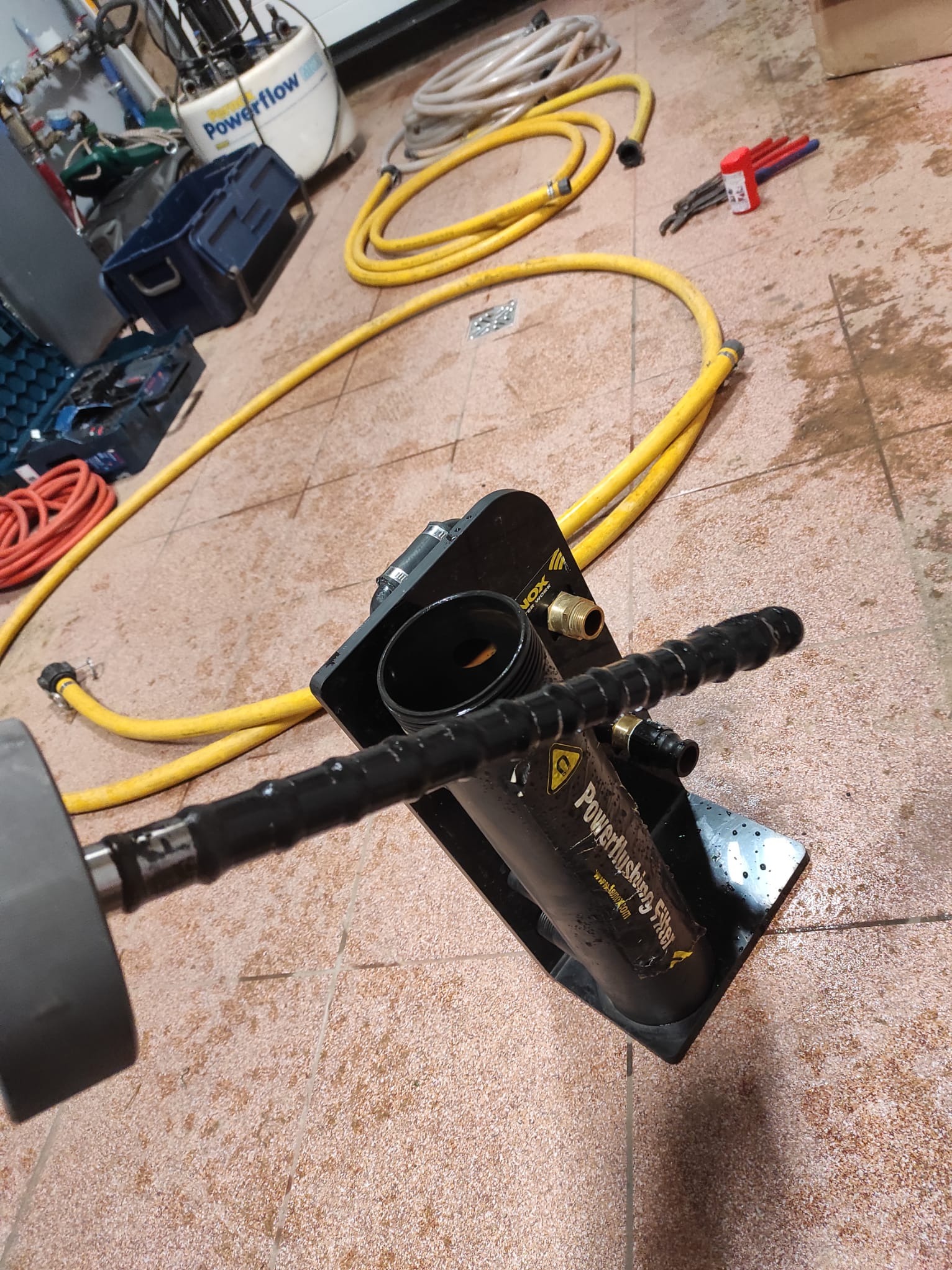 Urządzenie do płukania instalacji grzewczych Powerflushing Filter na tle żółtych węży i narzędzi na brązowej podłodze. Widok z góry, oświetlenie robocze.