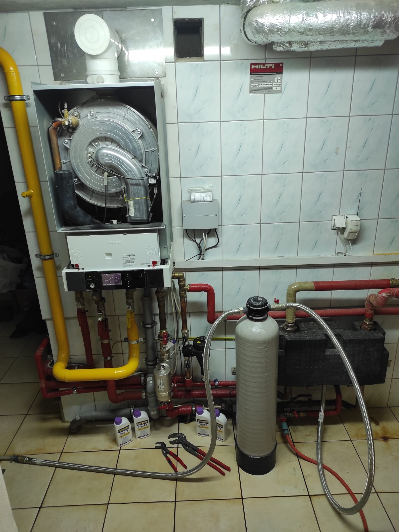 Montaż pieca Viessmann z filtrem wody i żółtymi rurami gazowymi na tle białych płytek. Widoczne narzędzia i preparaty czyszczące na podłodze.