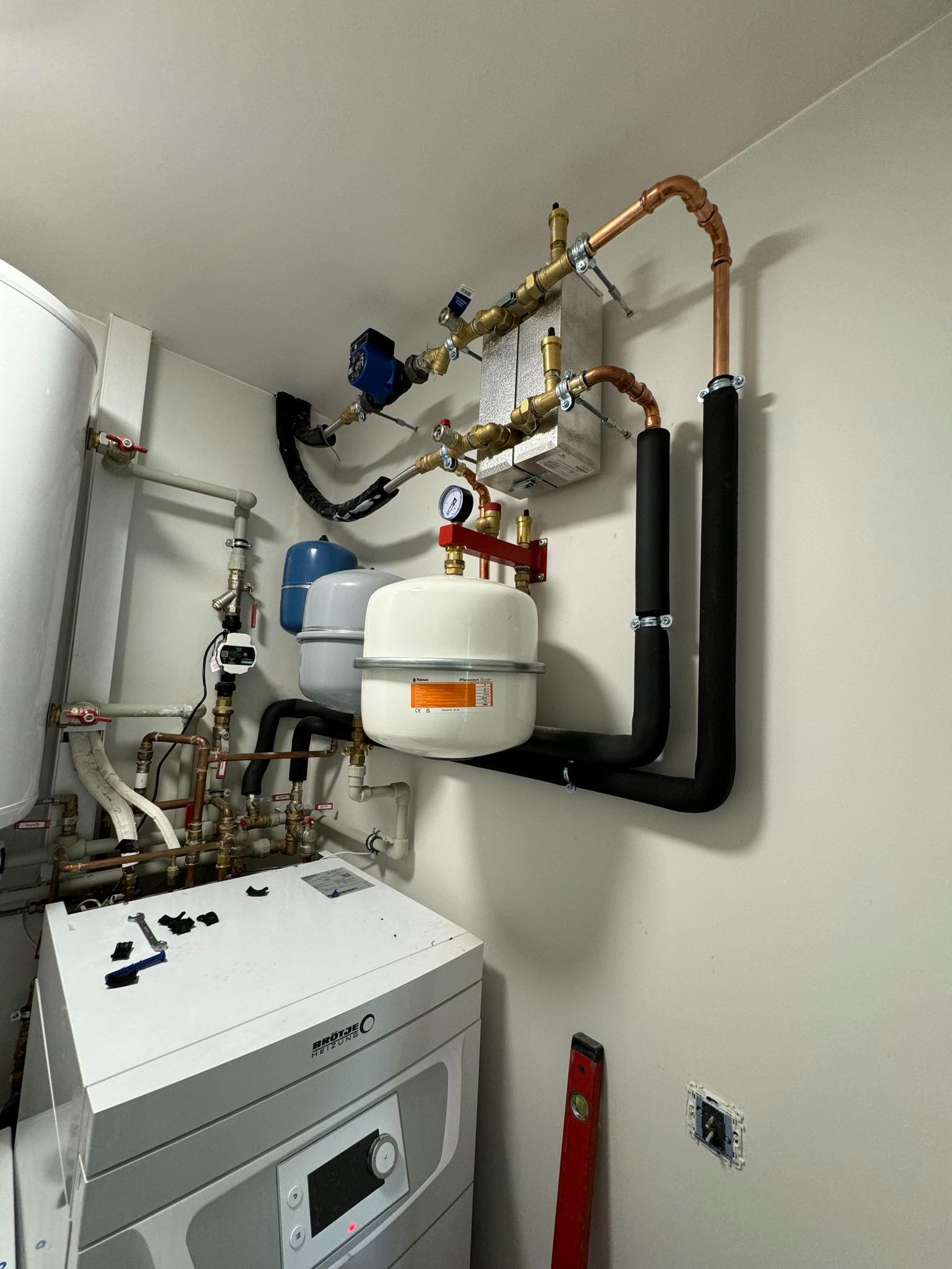 Złożona instalacja grzewcza w kotłowni: piec, zbiorniki wyrównawcze, rury miedziane z izolacją, zawory i manometr. Widoczny poziomica i narzędzia.