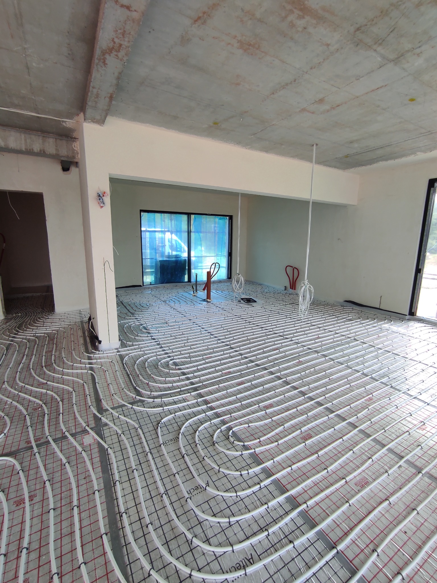 Białe rury ogrzewania podłogowego, ułożone na izolacji z siatką, w surowym wnętrzu z betonowym sufitem i dużymi oknami. Widok na rozległą instalację przed zalaniem posadzki.