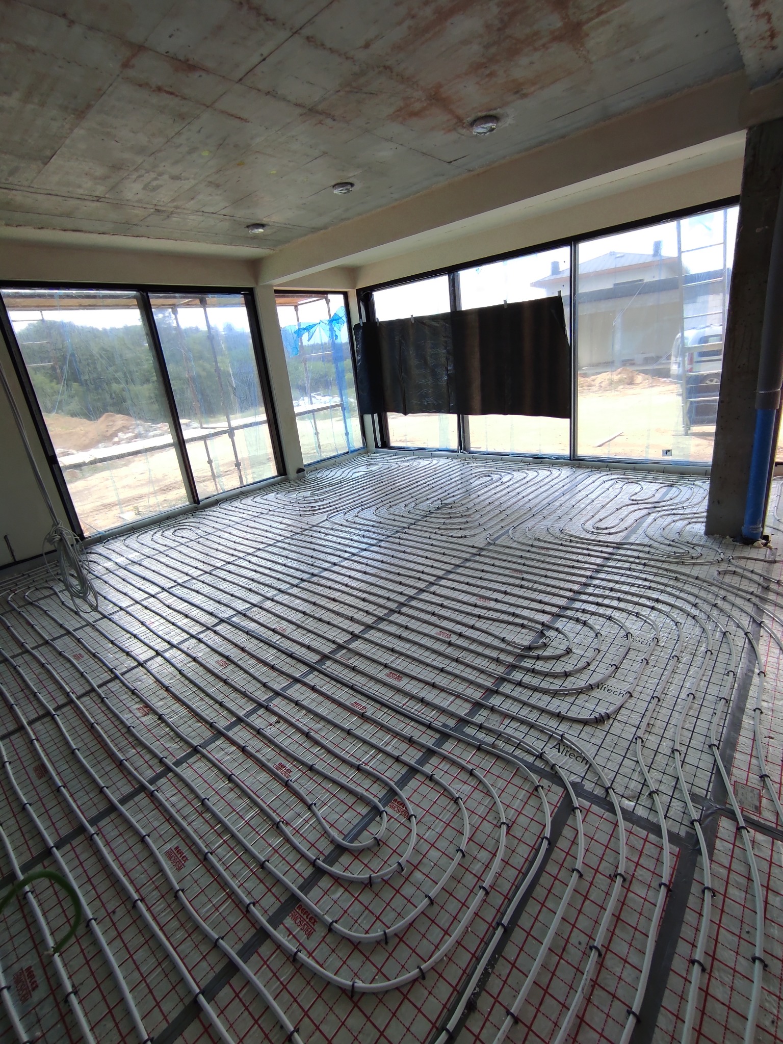 Instalacja ogrzewania podłogowego w surowym wnętrzu z dużymi oknami. Rury grzewcze ułożone na specjalnej siatce, widoczne złącza i fragmenty instalacji.