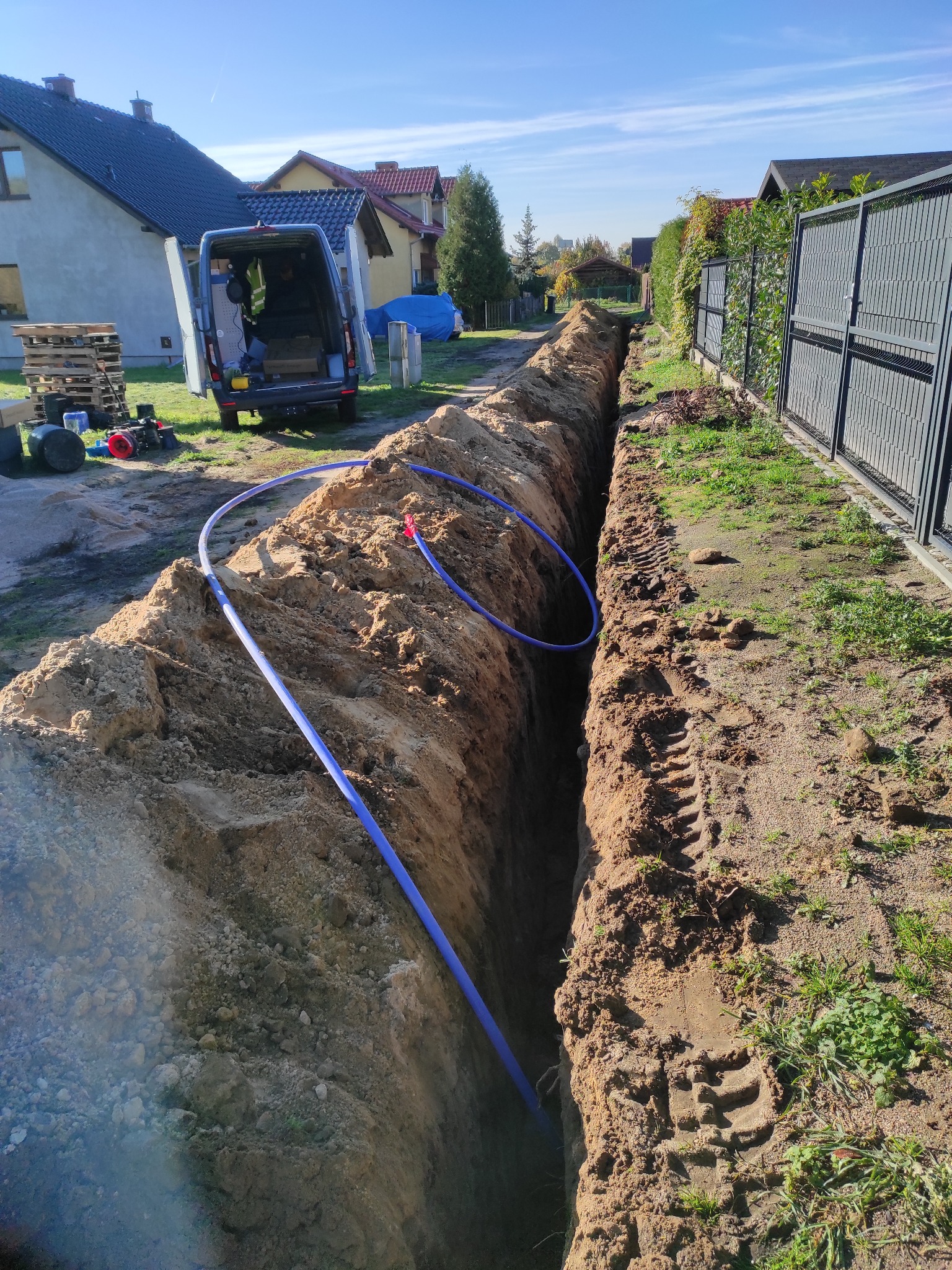 Wykop z niebieskim rurociągiem w ziemi, częściowo zasypany. W tle bus serwisowy, domy i błękitne niebo.