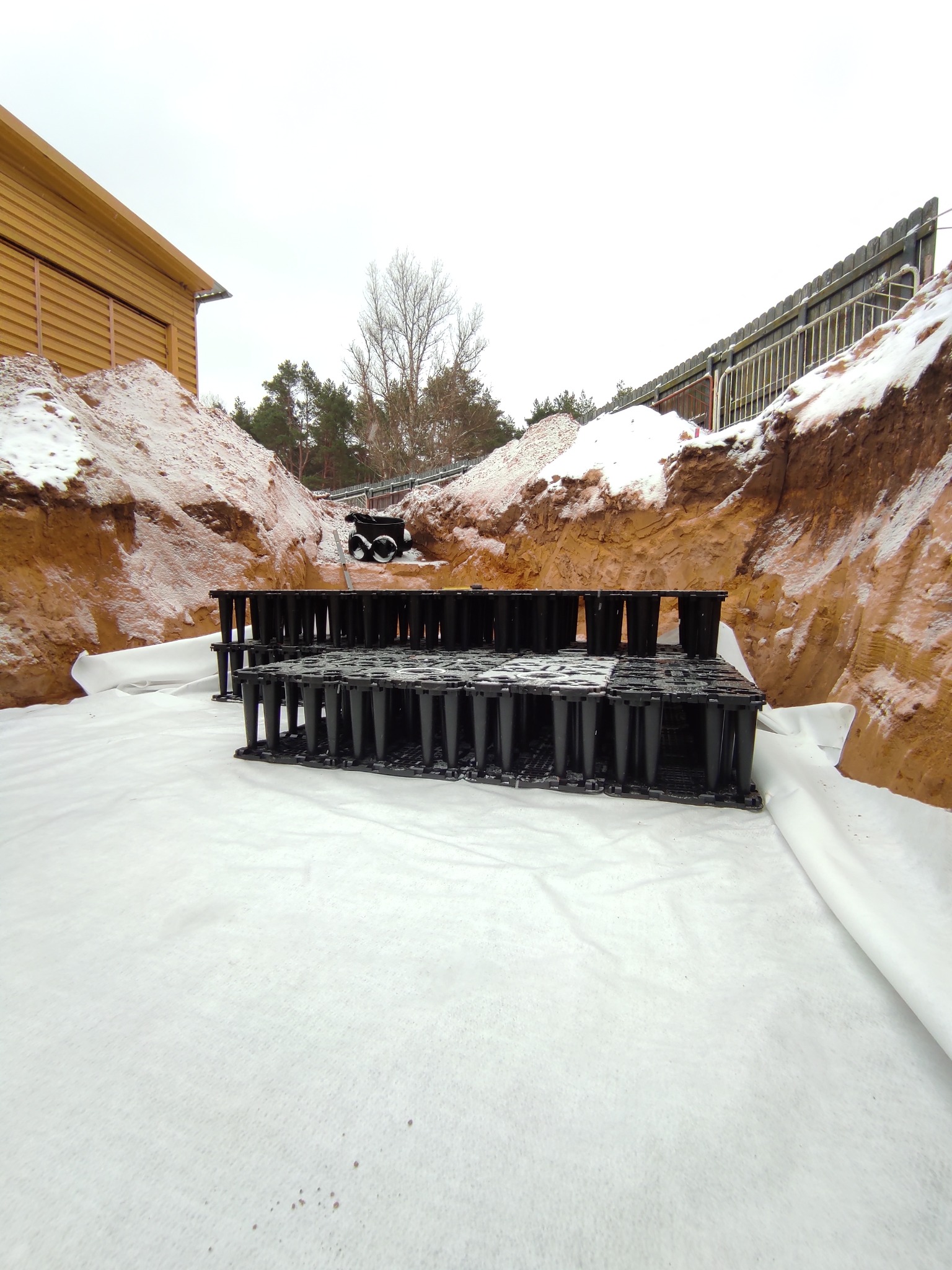 Instalacja drenażowa w wykopie, czarne moduły drenażowe ułożone na białej geowłókninie, ziemia z resztkami śniegu. Etap budowy przydomowej oczyszczalni ścieków.