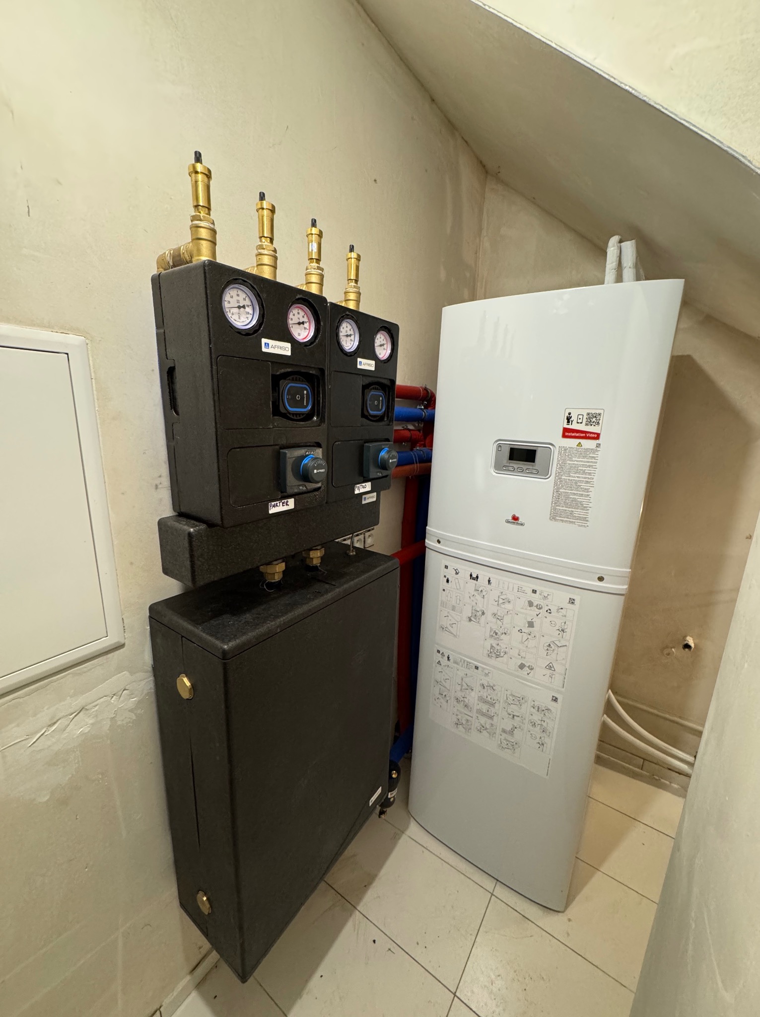 Zestaw Afriso z manometrami i zaworami, zainstalowany obok białego pieca grzewczego w pomieszczeniu z beżowymi ścianami i płytkami. Instalacja w skosie poddasza.
