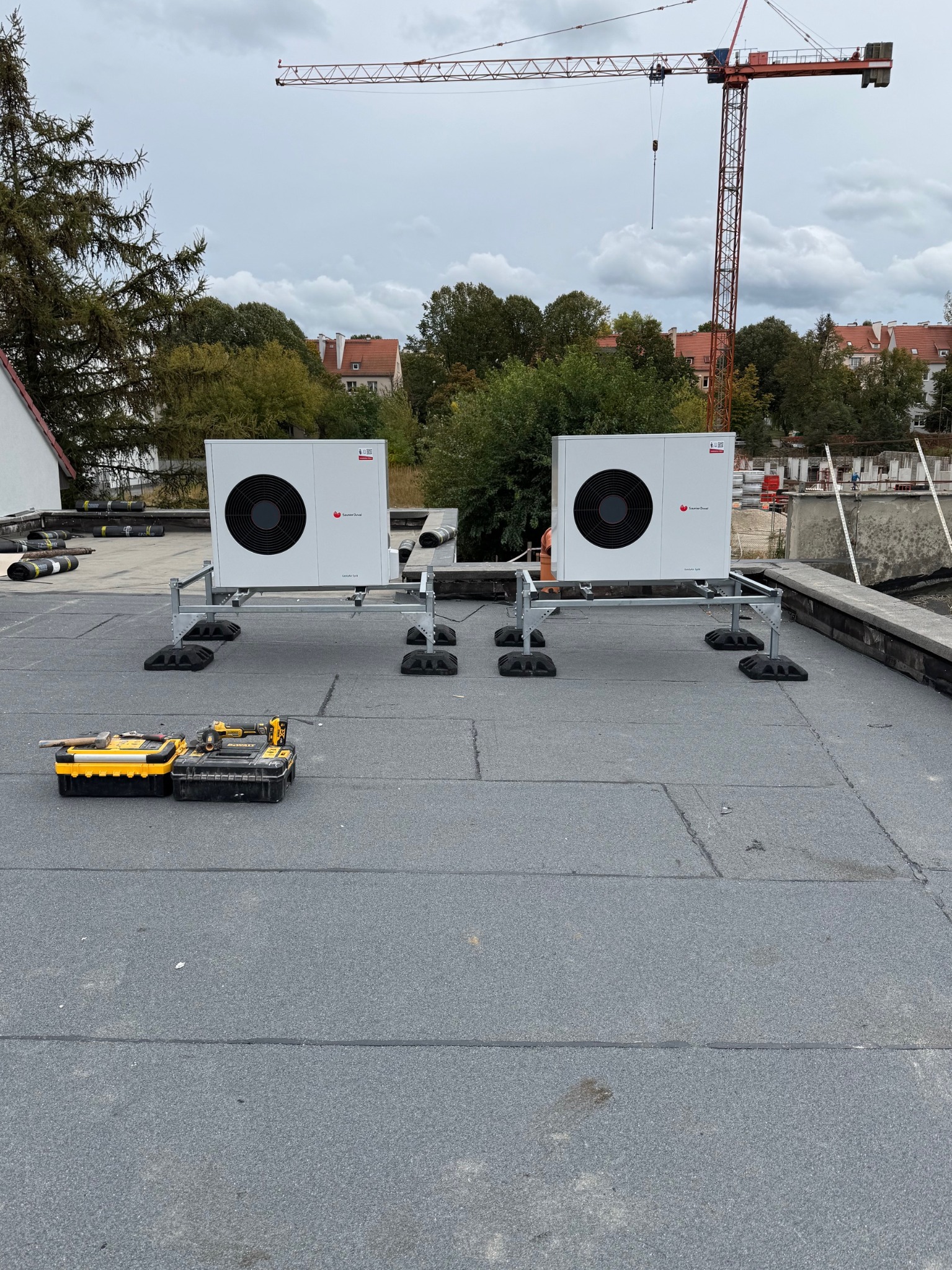 Dwie białe pompy ciepła na dachu budynku, osadzone na metalowych konstrukcjach. W tle widoczny żuraw budowlany i zabudowa miejska. Obok pomp leżą skrzynki z narzędziami.