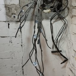 Delta - elektryk Gorzów - Przewody elektryczne wystające ze ściany z cegły i betonu, zabezpieczone taśmą z oznaczeniami, widoczne elementy instalacji w trakcie prac remontowych.