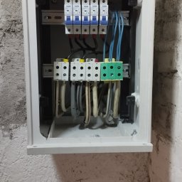 Delta - elektryk Gorzów - Otwarta, biała skrzynka elektryczna z bezpiecznikami i przewodami na szynach DIN, zamontowana na szorstkiej, pomalowanej ścianie. Widoczne złącza i kable w różnych kolorach.