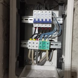 Delta - elektryk Gorzów - Otwarta skrzynka elektryczna z bezpiecznikami i przewodami na ścianie z surowego tynku. Widoczne elementy instalacji elektrycznej wewnątrz obudowy.