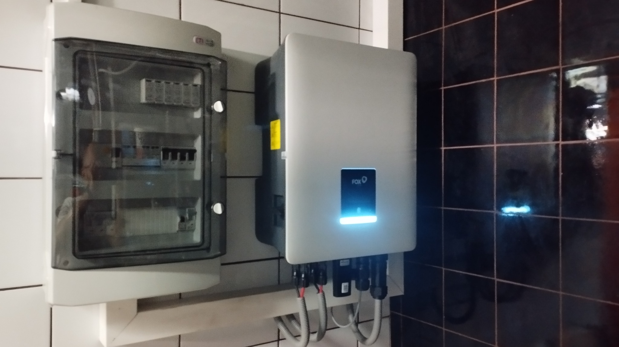 Szara szafka elektryczna z widocznymi bezpiecznikami obok białego inwertera z podświetlonym logo FOX, zamontowane na ścianie z białych i czarnych płytek.