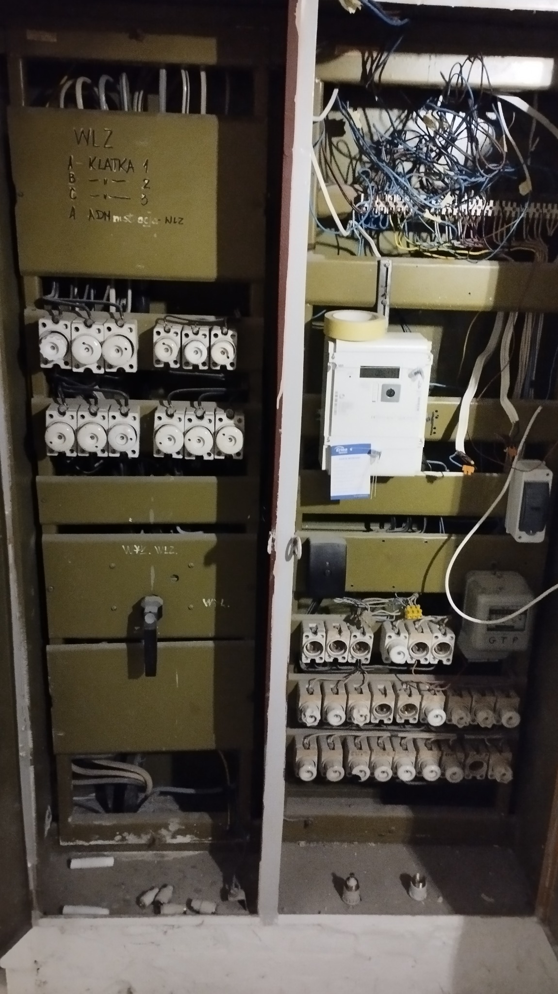 Otwarta, stara skrzynka elektryczna w odcieniach khaki z bezpiecznikami topikowymi, licznikiem energii i chaotyczną plątaniną przewodów. Widoczne napisy WLZ i oznaczenia klatek.