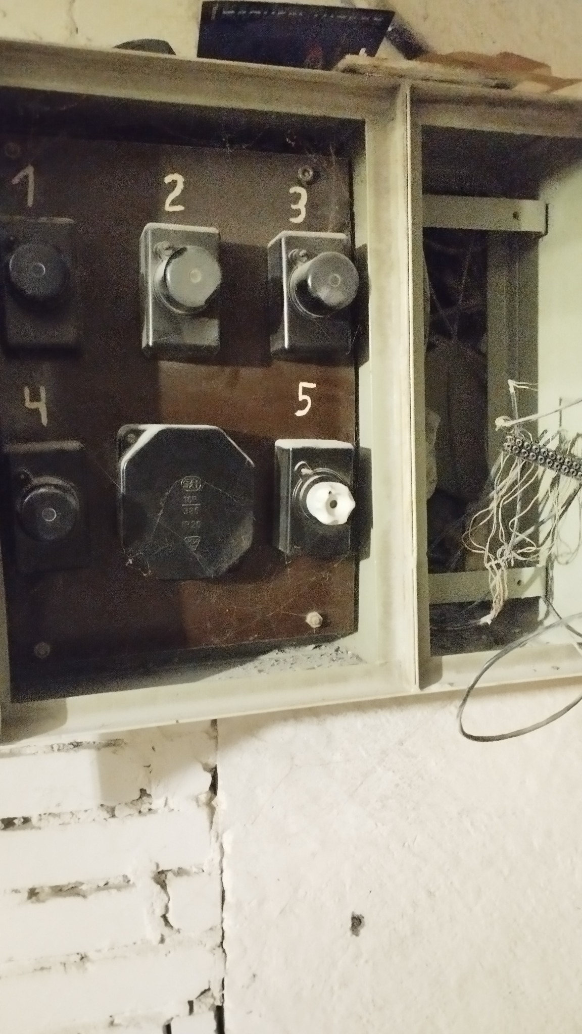 Stara, brudna skrzynka z bezpiecznikami z numerami 1-5, widoczne pajęczyny i zabrudzenia. Obok widoczne kable i przewody. Biała, pomalowana ściana z cegieł w tle.