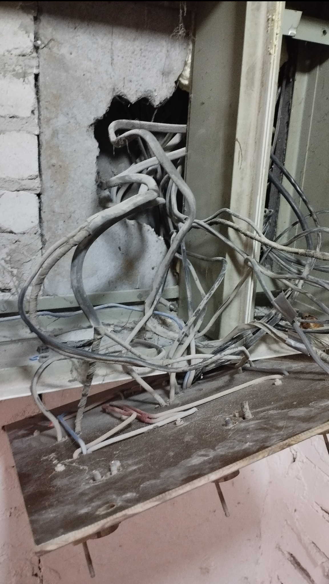 Skomplikowany układ kabli wychodzących z dziury w ścianie, widoczne przewody w szarych osłonach, fragment skrzynki elektrycznej. Niechlujne wykonanie.