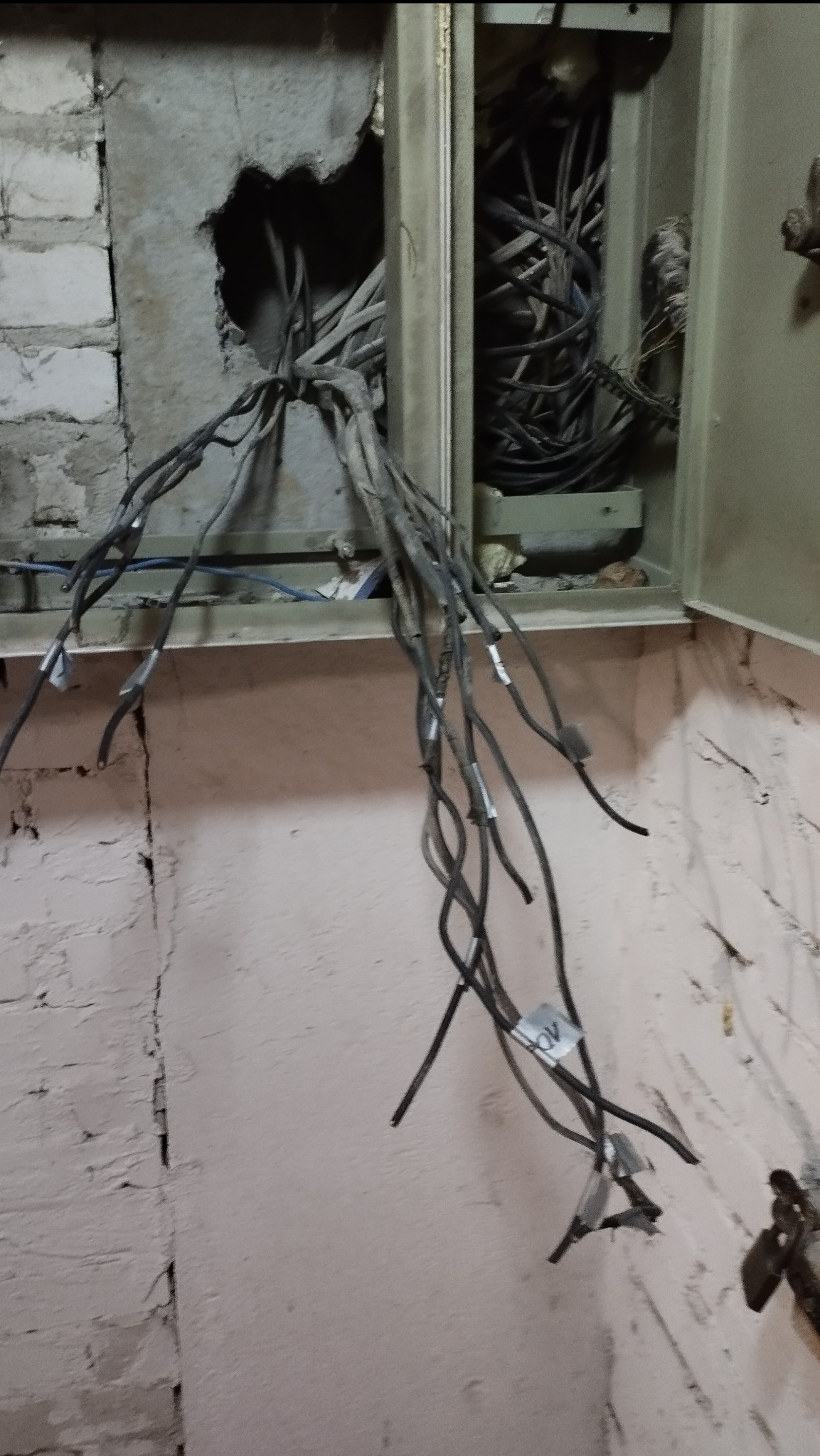 Otwarta skrzynka elektryczna z wystającymi przewodami, widoczne uszkodzenia ściany. Przewody oznaczone etykietami. Instalacja w trakcie prac.