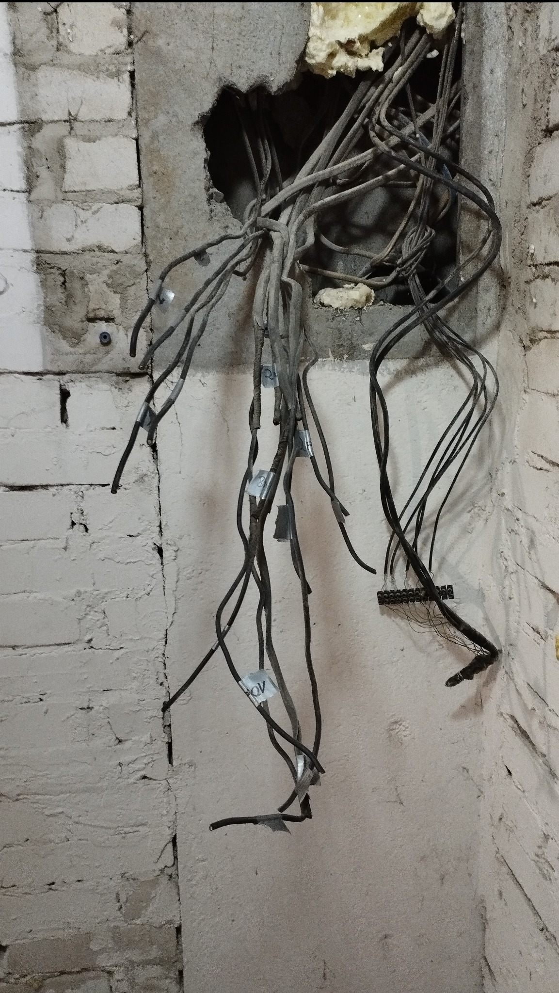 Przewody elektryczne wystające ze ściany z cegły i betonu, zabezpieczone taśmą z oznaczeniami, widoczne elementy instalacji w trakcie prac remontowych.