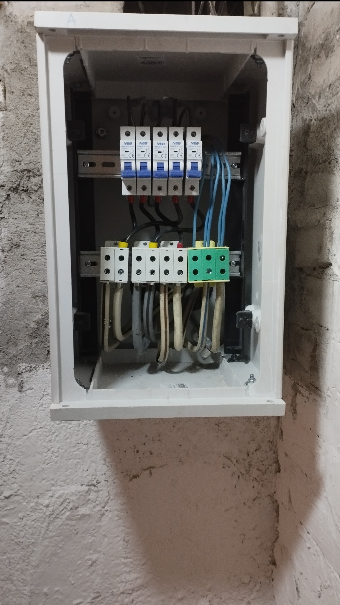 Otwarta, biała skrzynka elektryczna z bezpiecznikami i przewodami na szynach DIN, zamontowana na szorstkiej, pomalowanej ścianie. Widoczne złącza i kable w różnych kolorach.