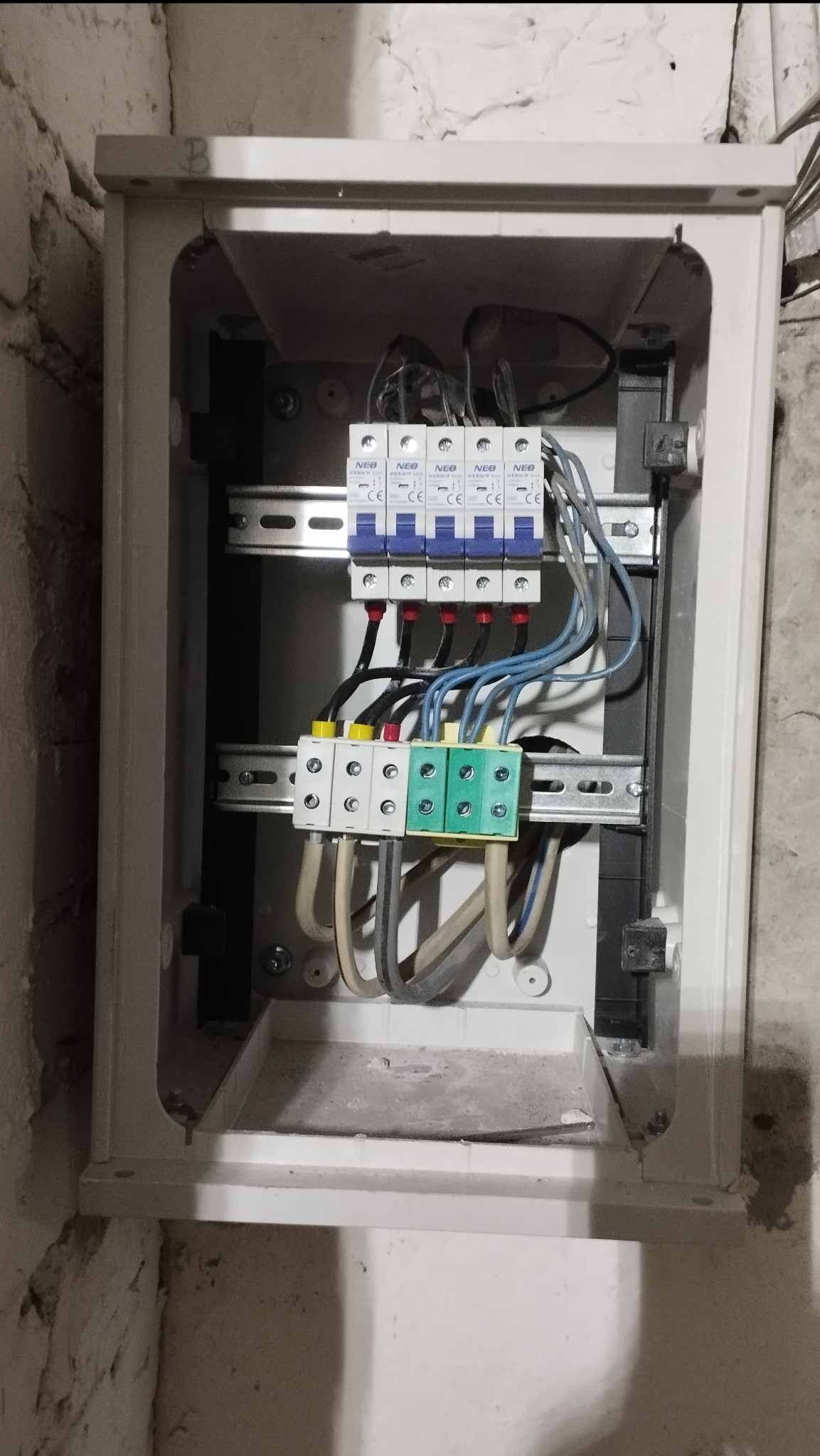 Otwarta skrzynka elektryczna z bezpiecznikami i przewodami na ścianie z surowego tynku. Widoczne elementy instalacji elektrycznej wewnątrz obudowy.