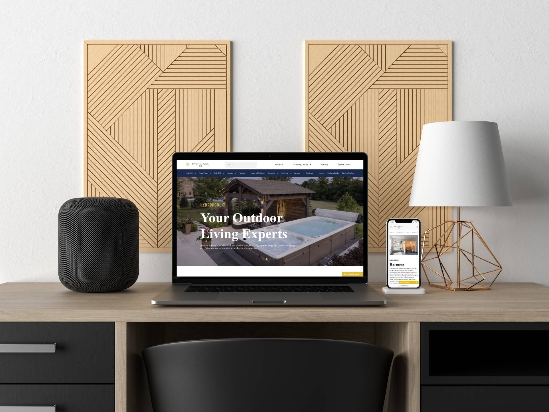 Laptop i smartfon wyświetlają stronę internetową firmy Hydropool z ofertą luksusowych swim spas, hot tubs i saun. Na biurku głośnik, lampka i dekoracyjne panele ścienne.