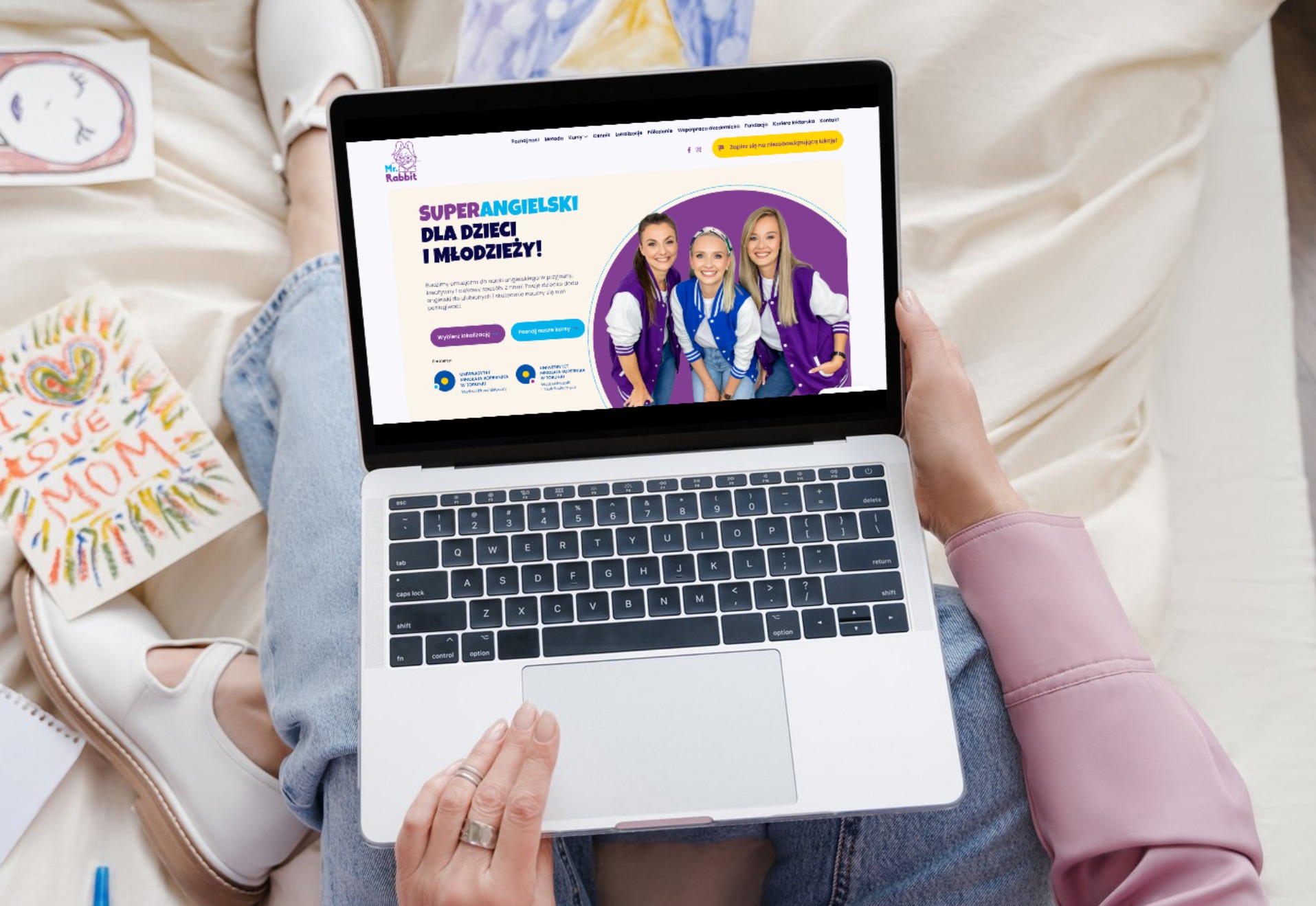Osoba siedząca na łóżku, trzymająca laptopa z wyświetloną stroną internetową szkoły językowej Mr.Rabbit dla dzieci i młodzieży, obok leżą rysunki dziecięce i notatnik.
