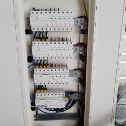 EcoDomes - Rozdzielnica elektryczna z zamontowanymi zabezpieczeniami marki Schneider, widoczne kable w kolorach niebieskim, żółtym i brązowym, oraz listwy zaciskowe w kolorze niebieskim i zielonym.