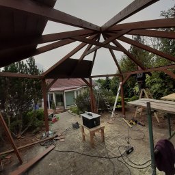 EcoDomes - Konstrukcja drewnianego tarasu w trakcie budowy, widok od wewnątrz, z widocznym pracownikiem na drabinie, narzędziami i częściami konstrukcji na brukowanym podłożu.