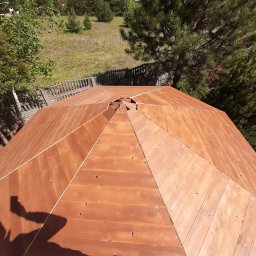 EcoDomes - Widok z góry na ośmiokątny, drewniany dach altany z widocznym zarysem osoby robiącej zdjęcie telefonem i fragmentem buta; w tle drzewa i trawnik.