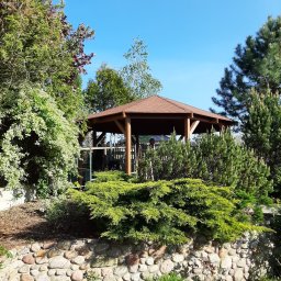 EcoDomes - Ośmiokątna altana ogrodowa z brązowym dachem, otoczona zielenią: kamienny murek, krzewy iglaste i liściaste, w tle błękitne niebo.