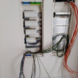 EcoDomes - Otwarta skrzynka rozdzielcza z widocznymi zabezpieczeniami, listwami zaciskowymi i przewodami w różnych kolorach, zamontowana na białej ścianie, z kalendarzem firmy Hager nad drugą skrzynką.