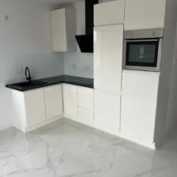 Nowoczesna, biała kuchnia z czarnym blatem i armaturą, błyszcząca podłoga imitująca marmur, zabudowa kuchenna z piekarnikiem i okapem.