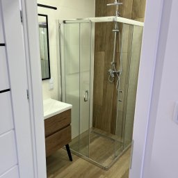 EcoDomes - Nowoczesna łazienka z kabiną prysznicową w rogu, drewnopodobne płytki ścienne i podłogowe, umywalka z szafką w kolorze drewna na czarnych nóżkach, widok zza częściowo otwartych białych drzwi.