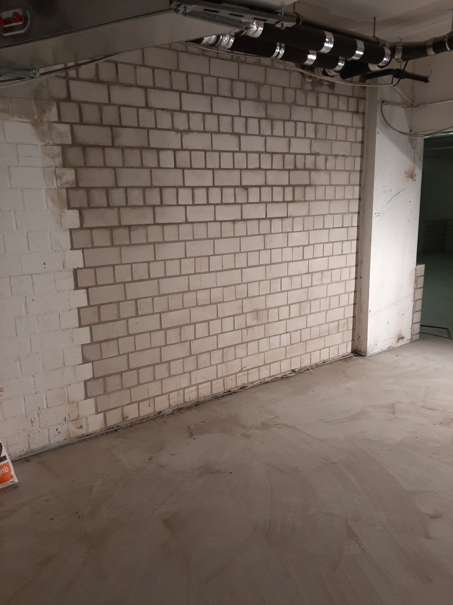 Częściowo pomalowana ściana z cegły w surowym wnętrzu, widoczne rury instalacji pod sufitem i przygotowane podłoże na podłodze.