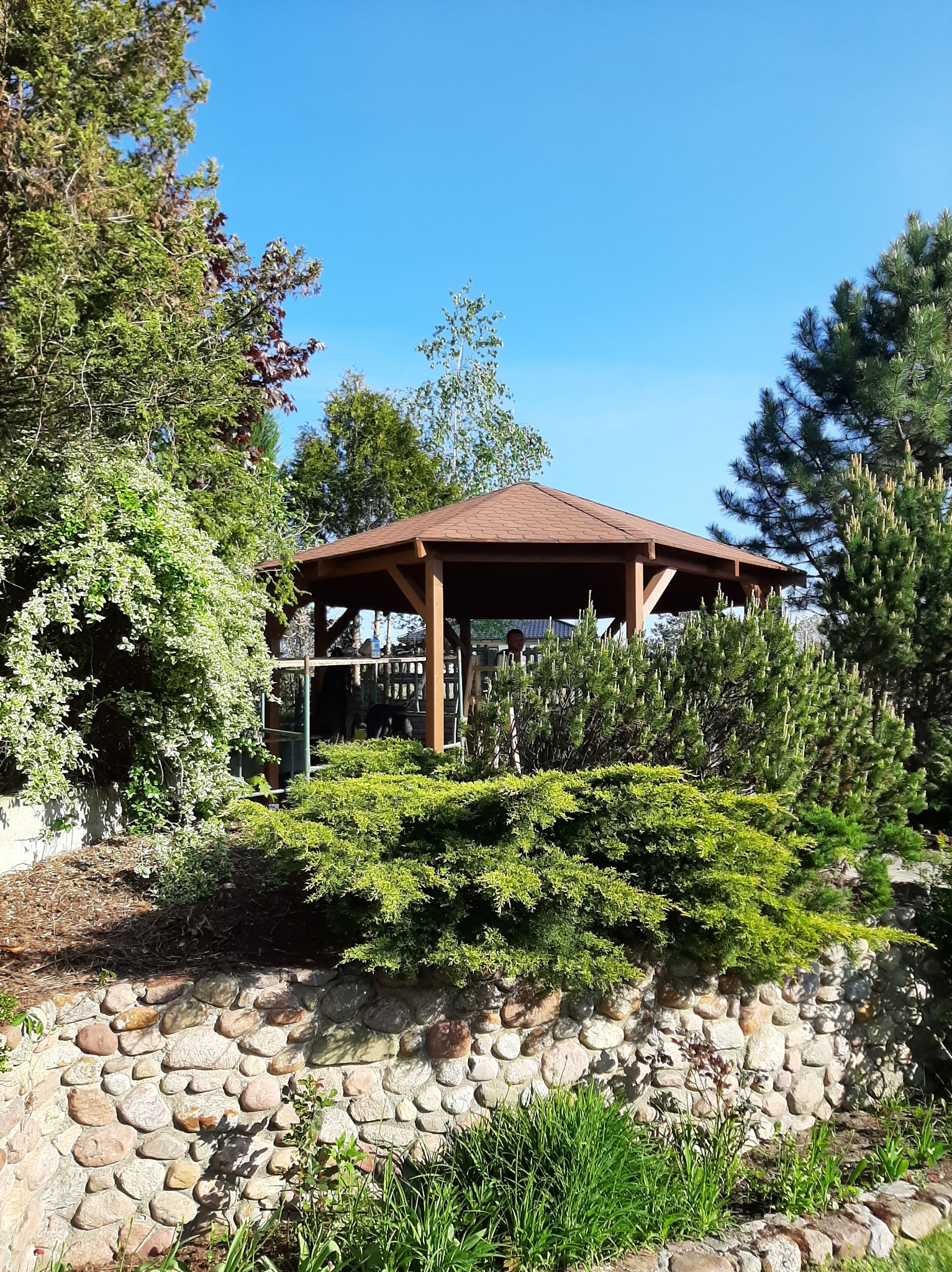 Ośmiokątna altana ogrodowa z brązowym dachem, otoczona zielenią: kamienny murek, krzewy iglaste i liściaste, w tle błękitne niebo.