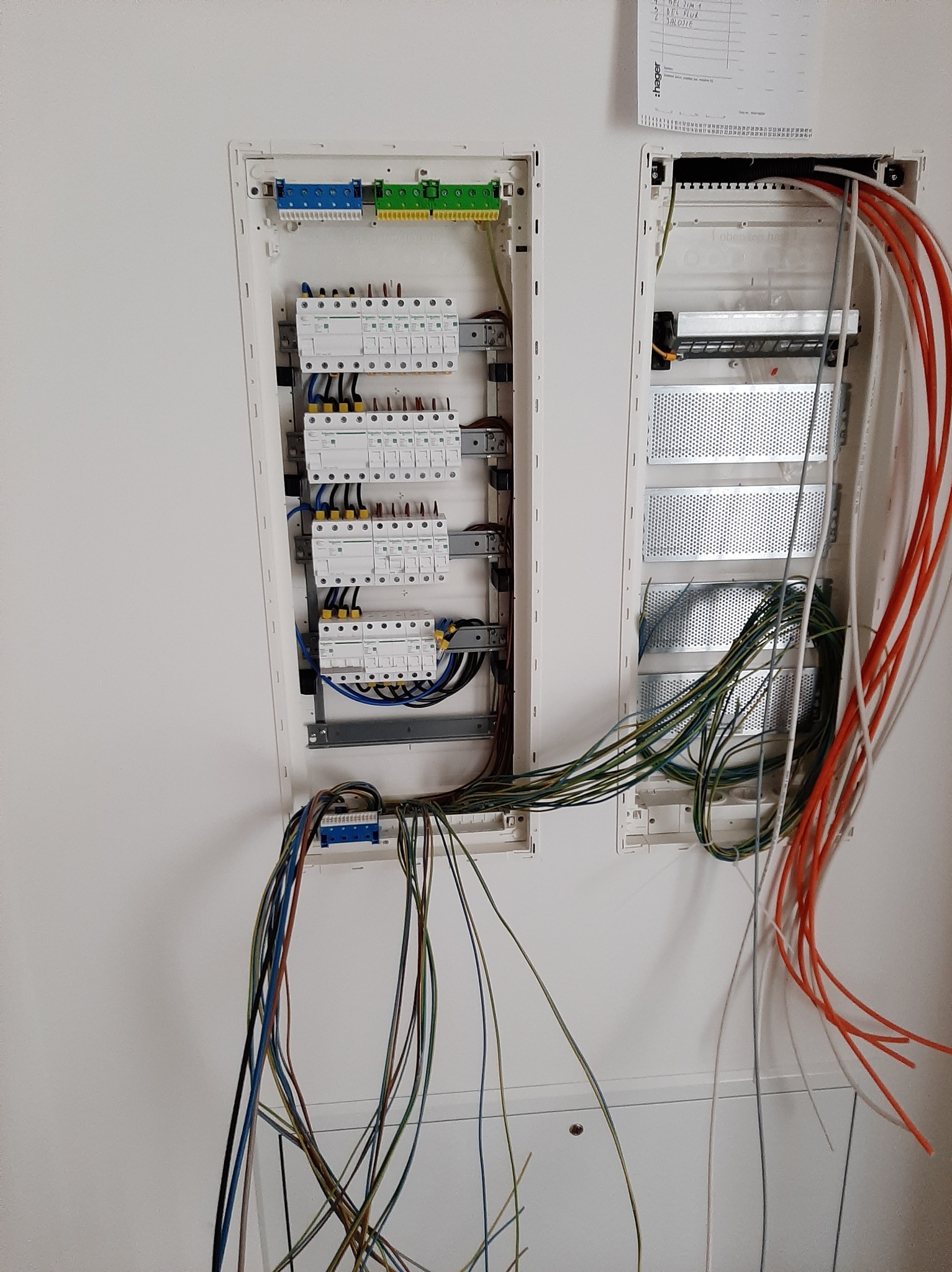 Otwarta skrzynka rozdzielcza z widocznymi zabezpieczeniami, listwami zaciskowymi i przewodami w różnych kolorach, zamontowana na białej ścianie, z kalendarzem firmy Hager nad drugą skrzynką.