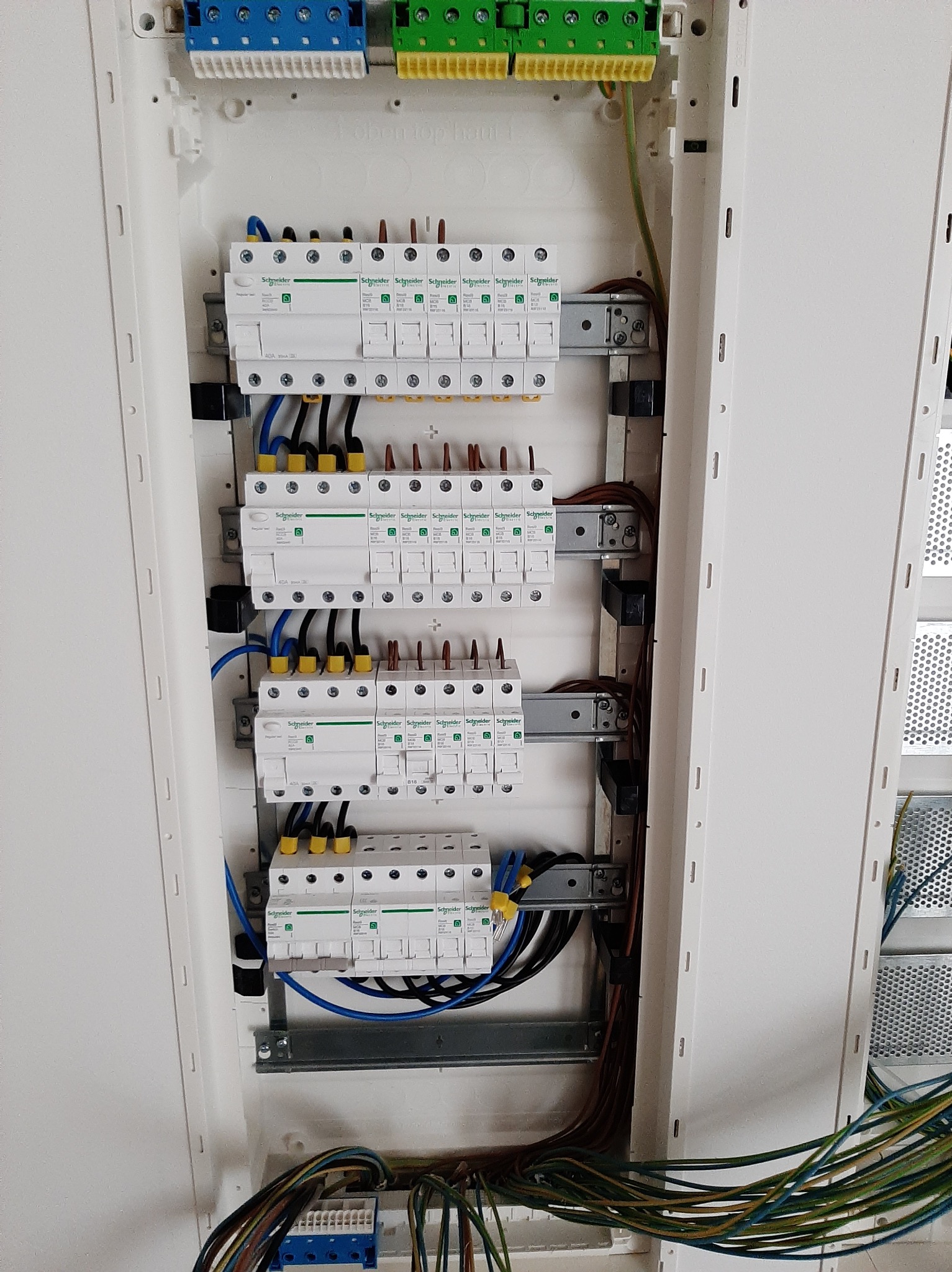 Rozdzielnica elektryczna z zainstalowanymi bezpiecznikami marki Schneider Electric, widoczne przewody w kolorach niebieskim, żółtym i brązowym, górne i dolne listwy zaciskowe z niebieskimi...