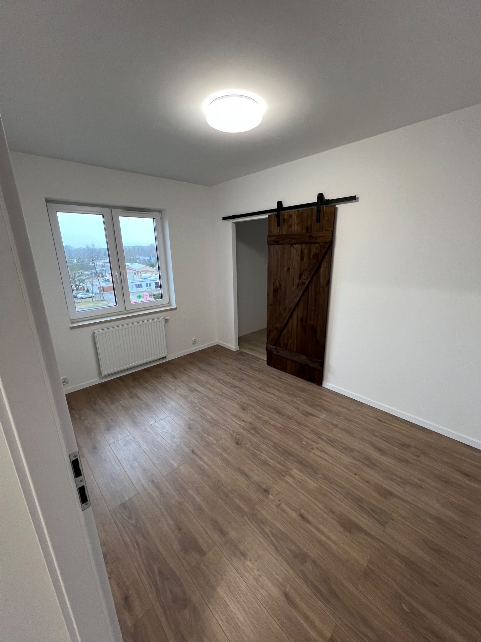 Jasne wnętrze pokoju z panelami podłogowymi, białymi ścianami, grzejnikiem pod oknem i przesuwnymi drzwiami stodołowymi z ciemnego drewna na czarnym torze.