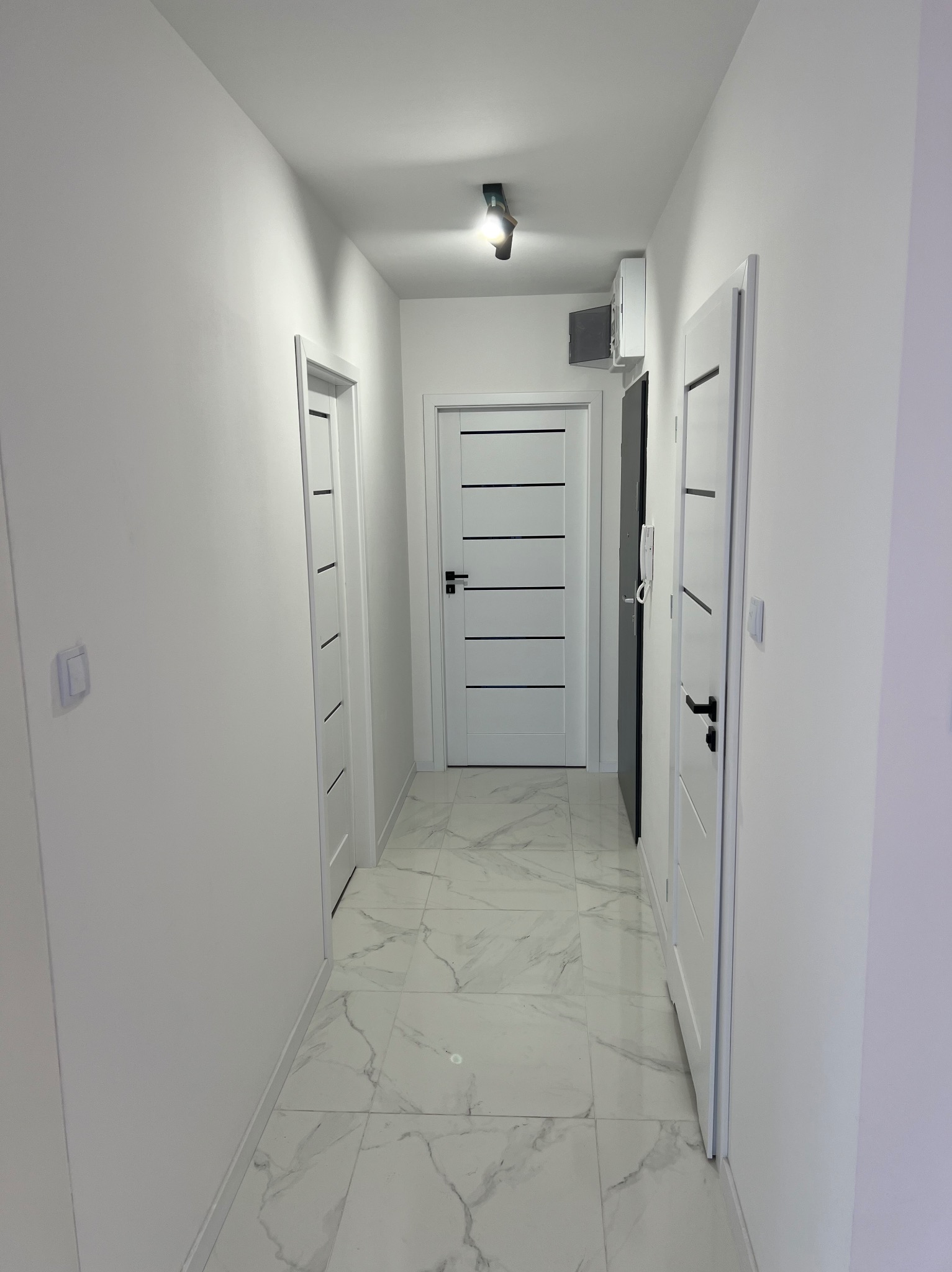 Biały korytarz z trzema parami białych drzwi z czarnymi poziomymi paskami, marmurowe płytki na podłodze, czarna lampa sufitowa i biała klimatyzacja na ścianie.