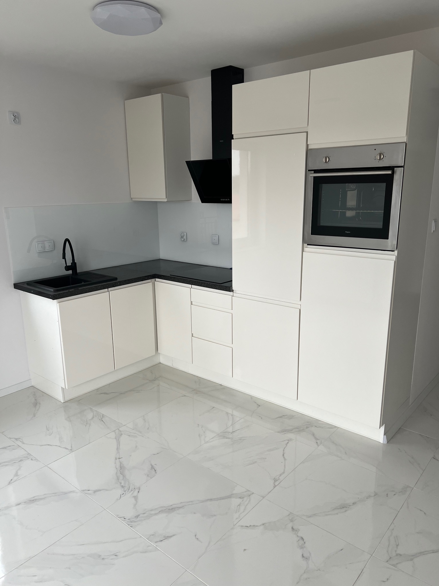 Nowoczesna, biała kuchnia z czarnym blatem i armaturą, błyszcząca podłoga imitująca marmur, zabudowa kuchenna z piekarnikiem i okapem.