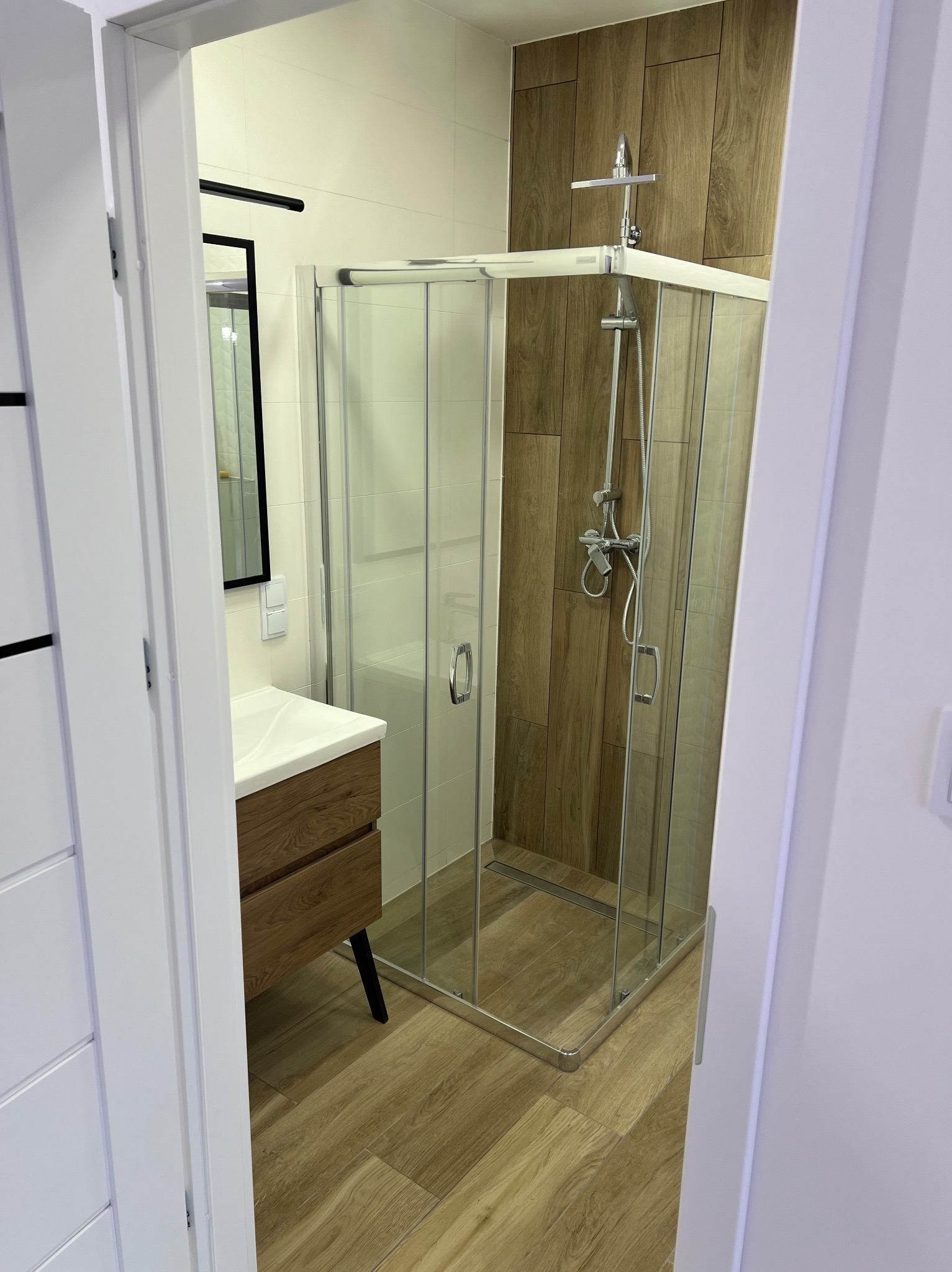 Nowoczesna łazienka z kabiną prysznicową w rogu, drewnopodobne płytki ścienne i podłogowe, umywalka z szafką w kolorze drewna na czarnych nóżkach, widok zza częściowo otwartych białych drzwi.