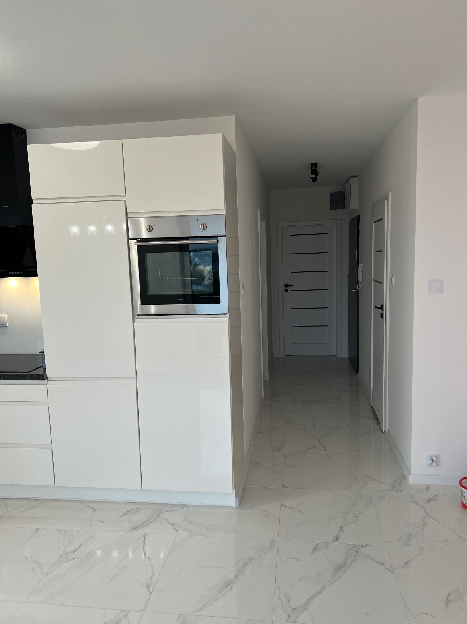 Nowoczesna, jasna kuchnia z białymi, lakierowanymi szafkami i wbudowanym piekarnikiem, widok na korytarz z marmurową podłogą i białymi drzwiami.