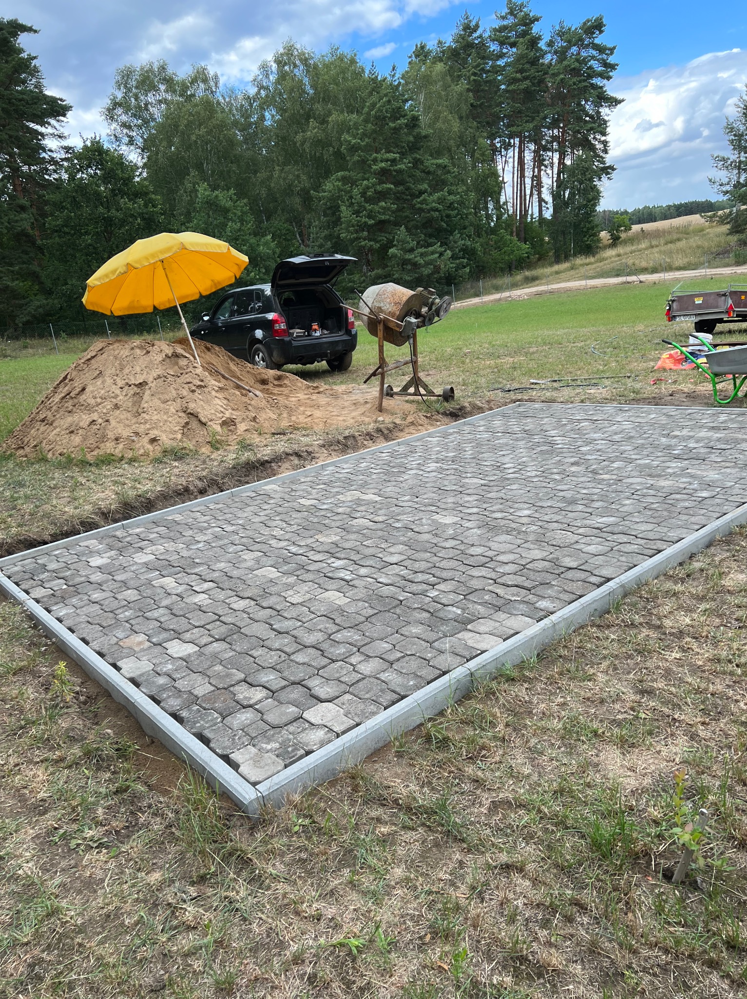 Przygotowany plac pod taras z kostki brukowej, obrzeża betonowe, w tle czarne auto z otwartym bagażnikiem, sterta piasku i żółty parasol.