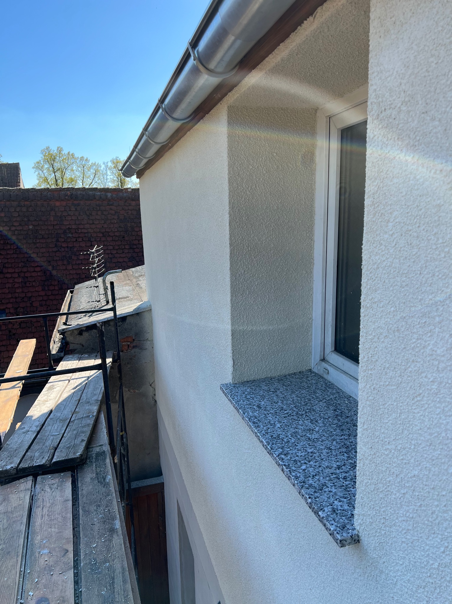 Elewacja budynku po szpachlowaniu i malowaniu, widok z rusztowania na okno z granitowym parapetem, rynna i antena na dachu sąsiedniego budynku.