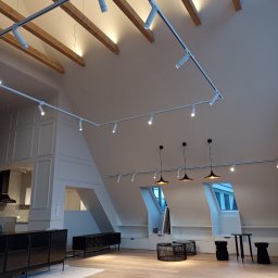 Andrzej Baldin metr.wow.pl - Nowoczesne poddasze po remoncie z drewnianą podłogą, białymi ścianami, drewnianymi belkami stropowymi, oświetleniem szynowym i wiszącymi lampami. W tle widoczna kuchnia.