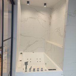 Andrzej Baldin metr.wow.pl - Nowoczesna, narożna wanna z hydromasażem, obudowana białą zabudową, z armaturą w chromowanym wykończeniu, na tle ściany wyłożonej białymi płytami imitującymi marmur, z widocznymi przewodami...