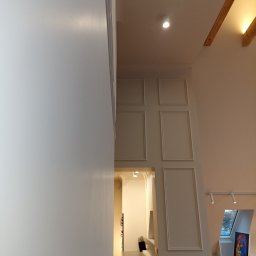 Andrzej Baldin metr.wow.pl - Wysokie pomieszczenie z białymi ścianami, sztukaterią i widokiem na kuchnię w głębi, oświetlone punktowo